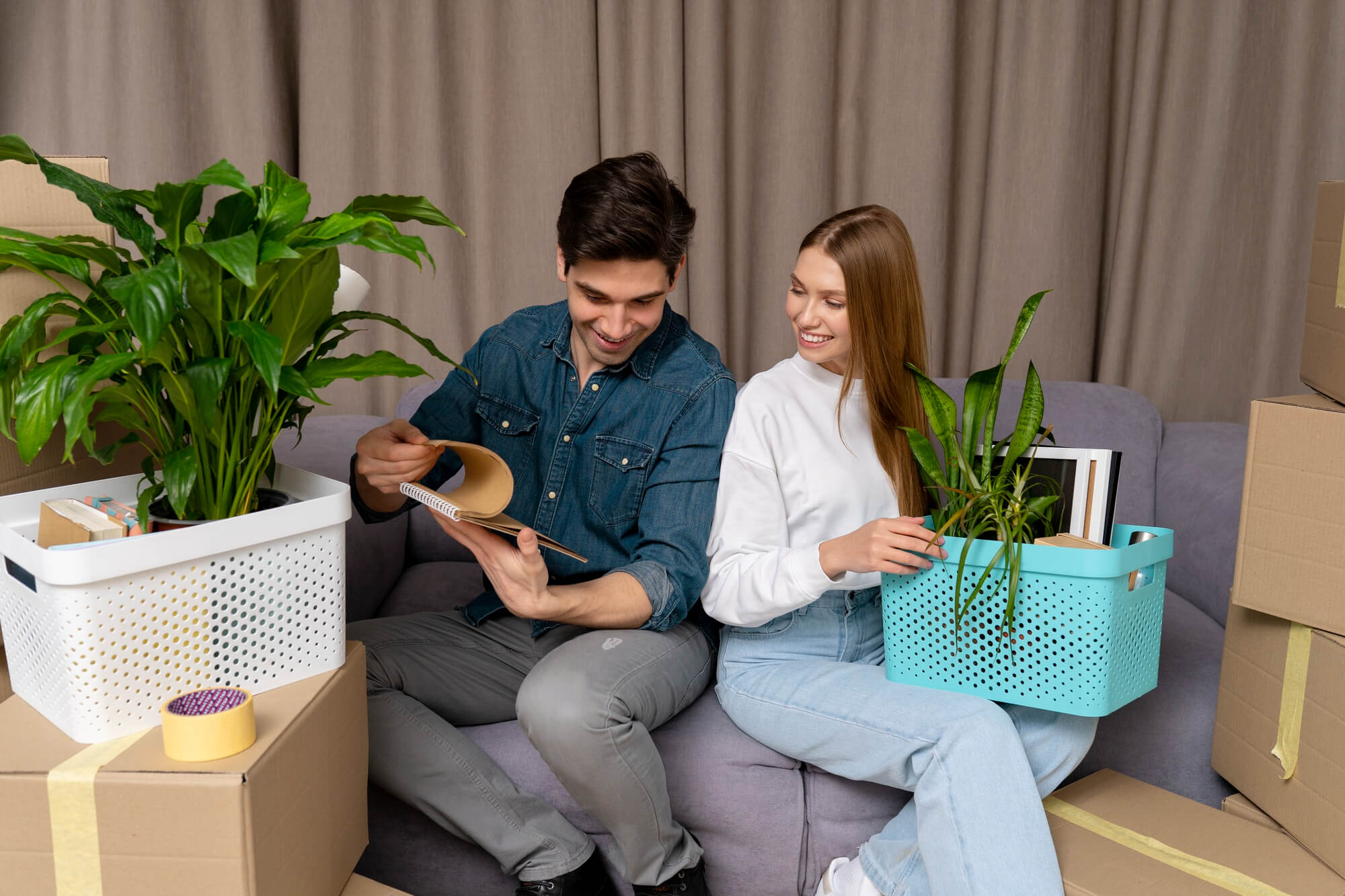 Smiling young couple on a gray sofa surrounded by moving boxes, the man holding a notebook and the woman holding a light-blue basket with a plant and books.