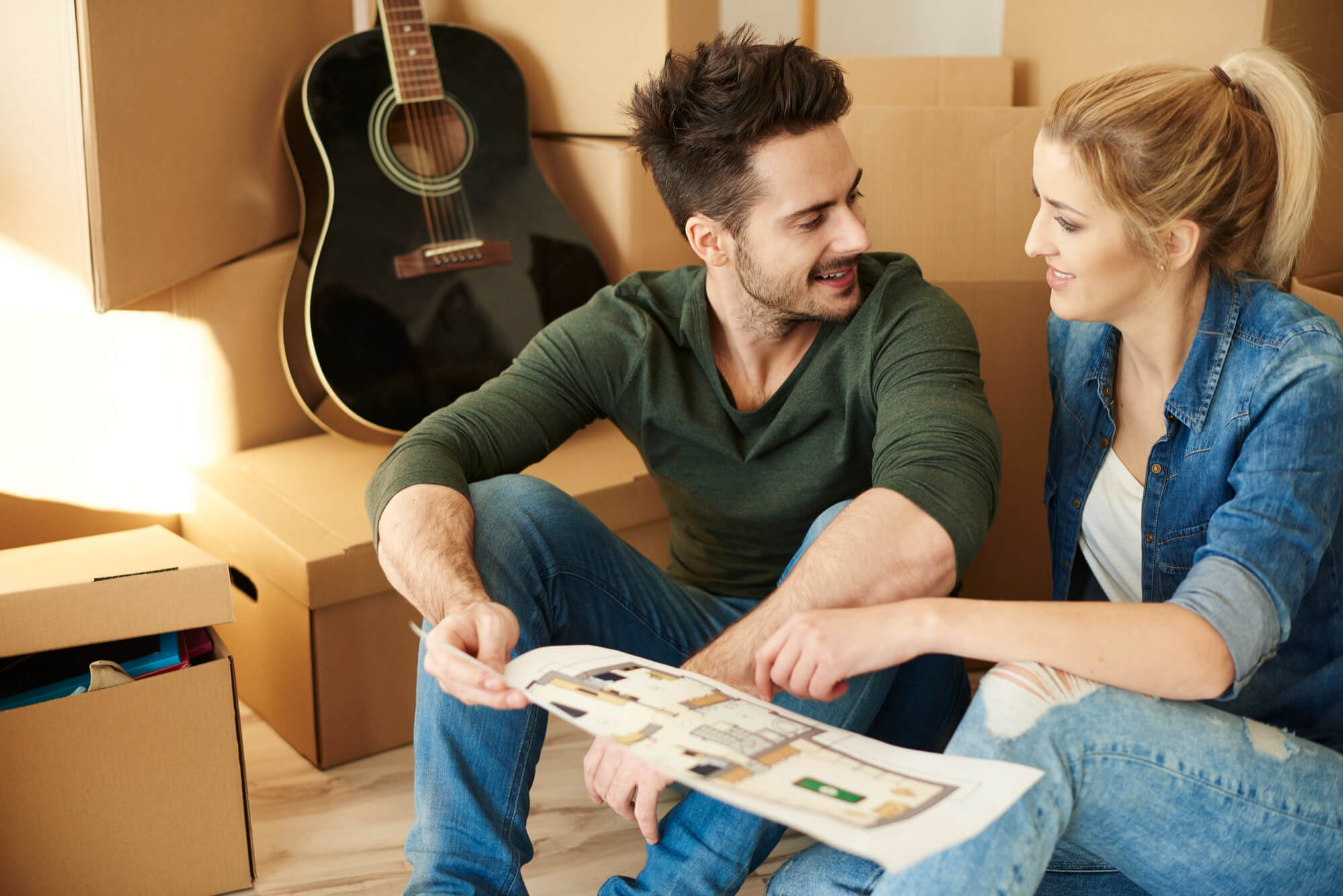 Smiling young couple sitting on the floor surrounded by cardboard boxes, reviewing a house floor plan together.